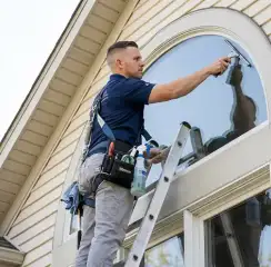 Professional window cleaner on a ladder using a squeegee to clean an exterior arched window, Overland Park, KS.